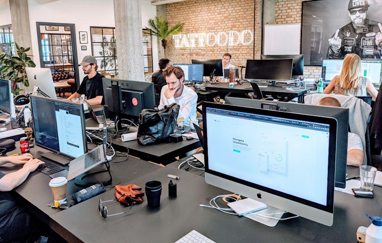 Developers at their desks in the open-plan office, with a Tattoodo neon sign on the brick wall